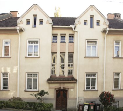 Zweistöckiges, helles Gebäude mit mehreren Fenstern und einem dekorativen Eingang.