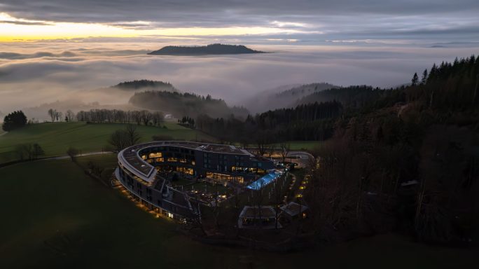 Landschaft mit Nebel über Wäldern und einem beleuchteten Stadion in der Dämmerung.
