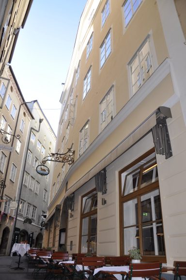 Blick auf ein Café mit Außenbestuhlung in einer schmalen Gasse mit hohen Gebäuden.